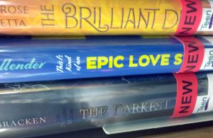 Three book spines, with the end of the titles obscured by stickers. The top one reads 'The Brilliant D-'. The middle one reads 'This is kind of an Epic Love S-'. The bottom one reads 'The Darkest Minds: The Darkest -'.